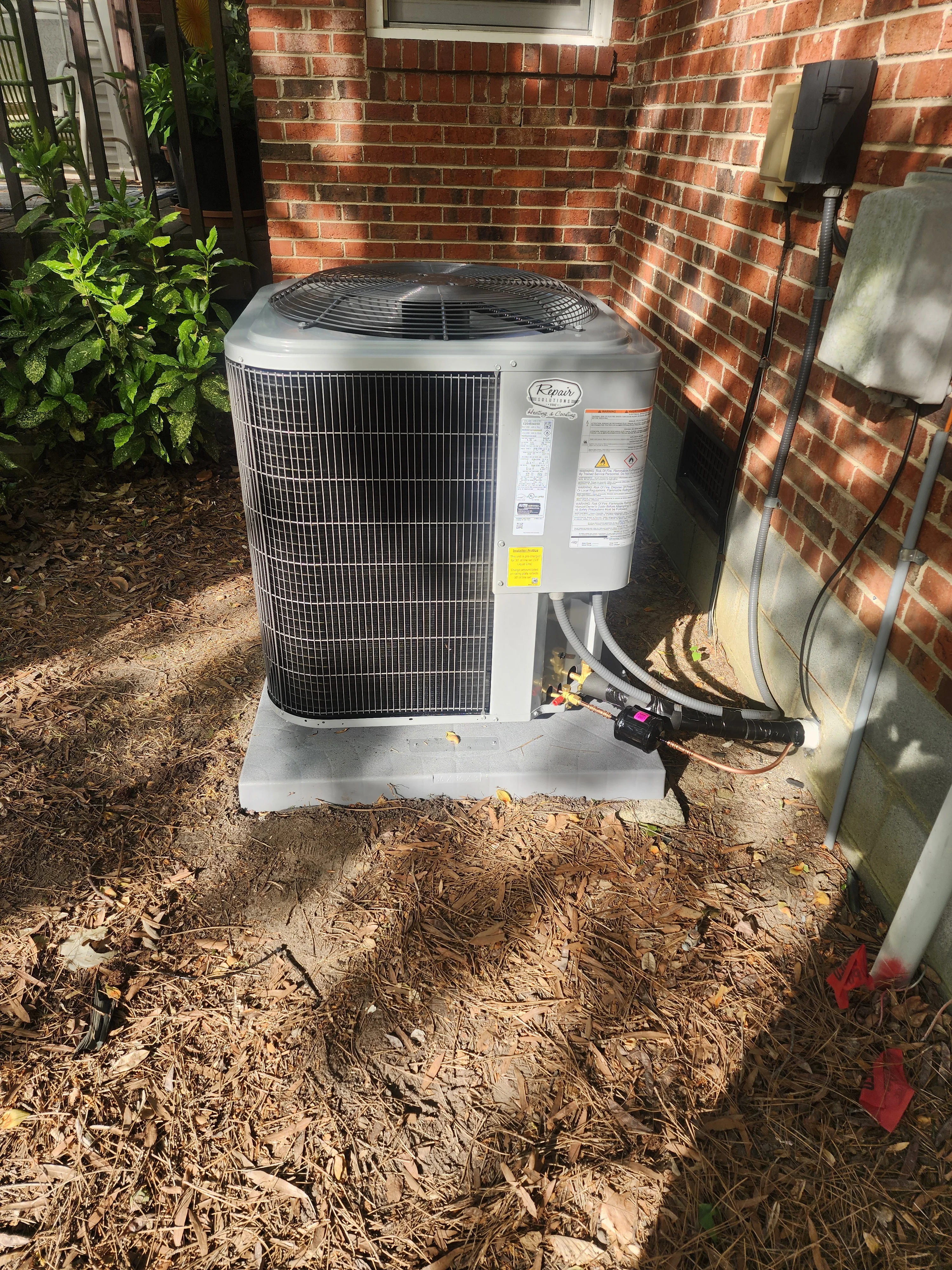 New AC condenser unit installed against brick wall with refrigerant lines and electrical connections